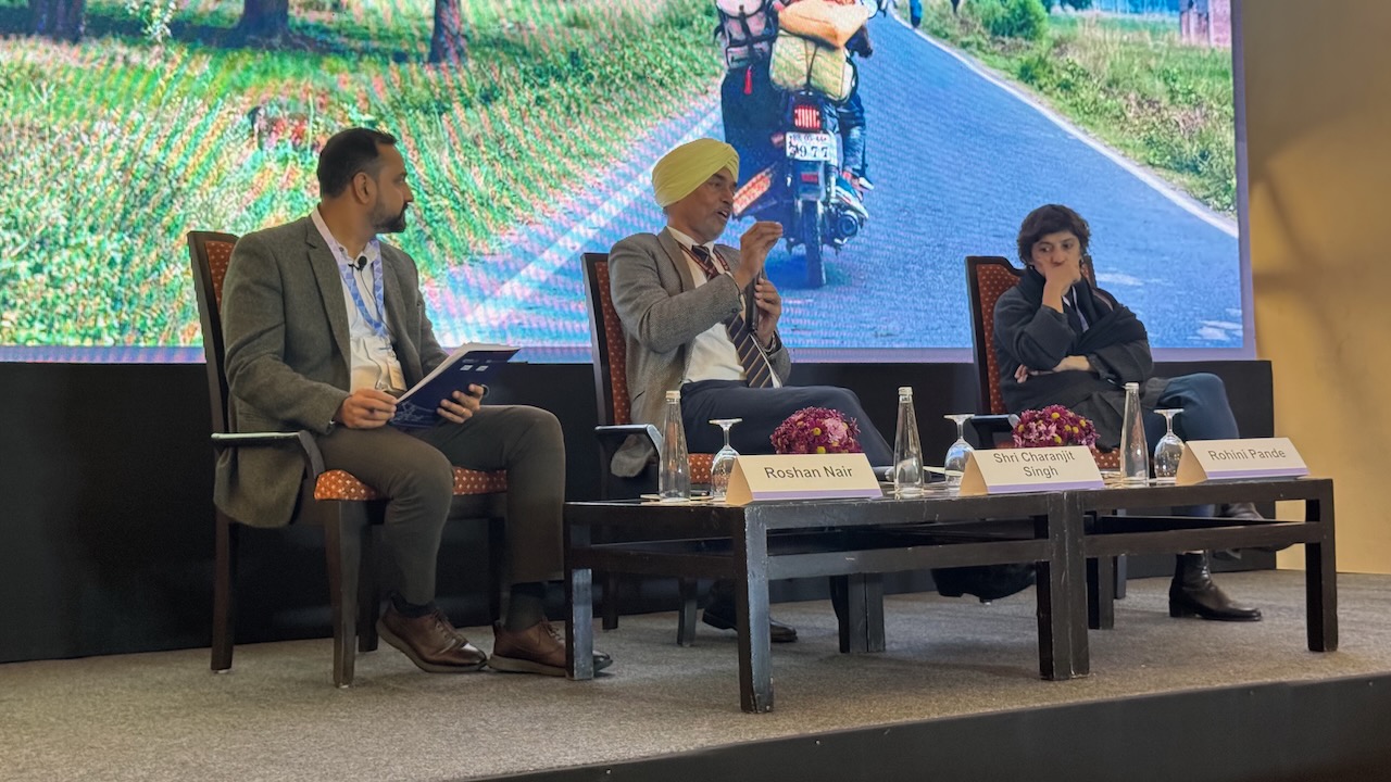 A man in a grey suit discusses policy issue with another man in a grey jacket and a woman in a grey jacket, seated on a stage in front of an audience